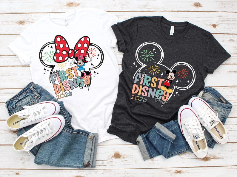 May include: Two t-shirts, one white and one charcoal gray, featuring a Mickey Mouse ear design with fireworks and the text "My First Disney Trip 2020." The shirts are paired with blue jeans and white sneakers.