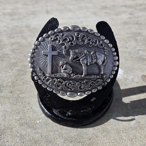 May include: Silver-colored oval belt buckle with a cross, a kneeling figure, and a horse with saddle. The buckle is set on a black horseshoe. The buckle has a detailed, ornate design with a beaded border.