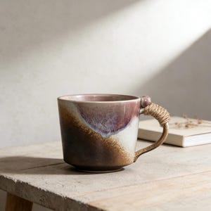 May include: A ceramic mug with a unique design. The mug features a handle wrapped in twine and a color gradient from dark brown to white and purple. The mug is sitting on a wooden surface.