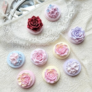 May include: A collection of nine circular decorative items, each topped with a sculpted floral design. The flowers include roses, peonies, and hydrangeas in shades of deep red, soft pink, lavender, pale yellow, and light blue on round bases.