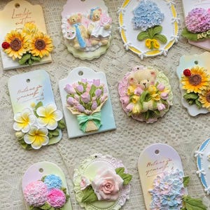 May include: A collection of decorative tags featuring 3D floral and teddy bear designs. The tags showcase sunflowers, tulips, hydrangeas, and roses in various colors. Some tags include text like "Love life and flowers" and "Believe in yourself."