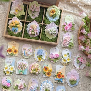 May include: A collection of decorative floral ornaments in various shapes and sizes. The ornaments feature detailed designs of sunflowers, roses, tulips, and other flowers, along with a teddy bear. The color palette includes pastels like pink, yellow, and blue.