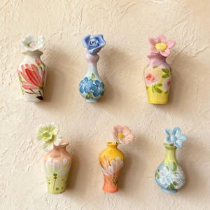May include: A collection of six small, decorative ceramic vases. Each vase is hand-painted with floral designs and topped with a matching flower. The vases feature a variety of colors, including pink, yellow, blue, and green.