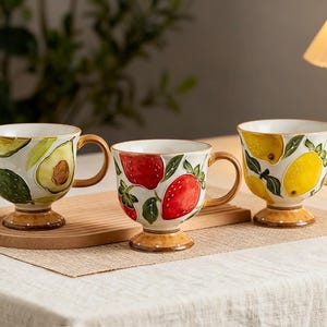 May include: Three ceramic mugs with fruit designs. One mug features avocado slices, another strawberries, and the third lemons. Each mug has a brown handle and base, and a white interior. The mugs are on a wooden tray.