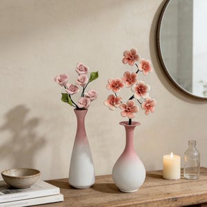 May include: Two ceramic vases with a white and pink ombre design hold artificial flowers. One vase has pink roses, the other has peach-colored blossoms. A small bowl, books, a candle, and a mirror are also on the wooden table.