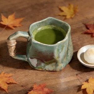 May include: A handmade ceramic mug with an irregular, organic shape and a glossy, mottled green and brown glaze. The handle is wrapped in natural jute twine. The mug is filled with a vibrant green matcha tea, showcasing a textured, earthy finish.