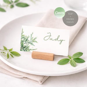 Pode incluir: Um cartão de lugar com o nome "Judy" em escrita verde, com vegetação em aquarela. O cartão repousa sobre um pequeno suporte de madeira, colocado em um prato branco com uma toalha de linho. O logotipo da Canva é visível.