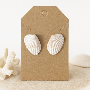 Puede incluir: Un par de pendientes de concha marina exhibidos en una etiqueta marrón. Los pendientes son blancos con sutiles detalles beige, imitando la textura natural de las conchas. El fondo incluye arena y coral.