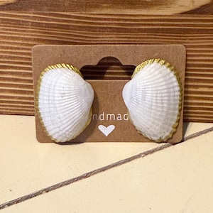 Puede incluir: Pendientes de concha blanca con borde dorado, presentados en una tarjeta marrón con la palabra "ndmad" y un símbolo de corazón. Fondo de madera.