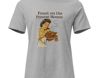 May include: A light gray t-shirt featuring a retro illustration of a woman holding a roasted turkey. The text "Feast on the Present Moment" is above the illustration in brown letters. The woman is wearing a yellow shirt and white apron.