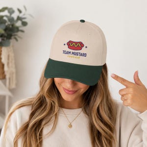 May include: A two-tone baseball cap with a cream-colored crown and a dark green brim. The front features a graphic of a hot dog with yellow mustard, flanked by two small stars, and the text "TEAM MUSTARD FOREVER" printed in dark blue and yellow.