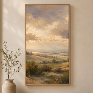 May include: A vertical framed landscape painting depicting a rolling countryside at sunset. The scene features golden light illuminating fields, scattered trees, and distant hills under a cloudy sky with soft shades of beige, brown, and muted blue.