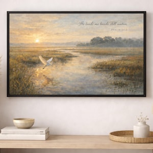 May include: A framed landscape painting shows a marsh scene at sunset. A white bird flies over the water, reflecting the golden light. The text "He leads me beside still waters. Psalm 23:2" is written in the upper right corner.