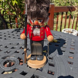 May include: A wooden nutcracker figure dressed as a British guardsman with a tall, dark brown faux fur bearskin hat, a bright red jacket with gold buttons, and black trousers. The figure holds a tall black rifle and stands on a round base.