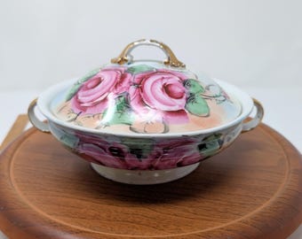 May include: A covered ceramic serving bowl with a floral rose design. The bowl has a white base with pink, red, and green roses. The lid has a gold handle. The bowl is sitting on a wooden tray.