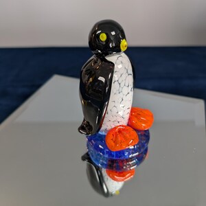 May include: A glass penguin figurine with a black head, white and black body, yellow beak, and orange feet. The penguin stands on a blue and orange base, reflecting on a mirrored surface. The figurine is a decorative item.