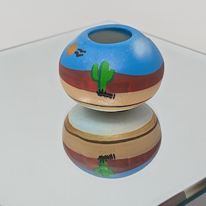 May include: A small, round, ceramic pot featuring a hand-painted desert landscape. The design includes a bright blue sky with a yellow sun, a dark red horizon line, and a green cactus set against a tan base. The pot has a smooth, matte finish.