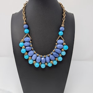 May include: A statement necklace with a gold-tone chain and a bib of blue and turquoise beads. The necklace is displayed on a black mannequin. The beads are oval and round, creating a layered effect.