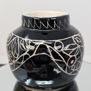 May include: A small, round, black ceramic vase with a glossy finish. The exterior features hand-carved white line art depicting a continuous botanical pattern of leaves and berries, with a zigzag pattern encircling the neck.