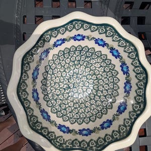 May include: A round ceramic bowl with a scalloped edge, featuring a cream-colored base decorated with a repeating dark green geometric pattern and a border of blue floral motifs with green leaves, viewed from directly above in a top-down perspective.