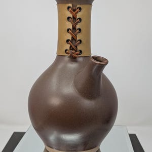May include: A dark brown ceramic vessel with a rounded body and a narrow neck. The neck is wrapped in a tan leather band secured with dark brown leather lacing through black metal eyelets. A small, angled spout extends from the side of the body.