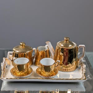 May include: A miniature gold-colored tea set on a white tray. The set includes a teapot, sugar bowl, creamer, and two teacups with saucers. The tea set has a shiny, reflective surface and is likely made of ceramic.