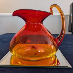 May include: A vibrant, hand-blown glass pitcher with a curved handle. The pitcher transitions from a deep red at the top to a warm amber at the base. It sits on a mirrored surface, reflecting its form and colors. A decorative piece for serving beverages.