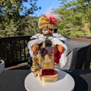 May include: A wooden nutcracker figure representing Balthasar, wearing a gold turban with a red feather, a white faux fur-trimmed cape, and a gold sash. The figure holds a gold-colored incense burner and stands on a base labeled C+M+B BALTHASAR.