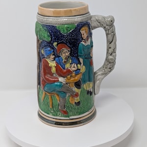 May include: A ceramic beer stein with a detailed relief scene of people gathered around a table. The stein features a gray handle, a tan rim, and a green, blue, and brown color scheme. The design includes figures in traditional clothing.