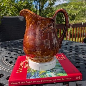 May include: A brown glazed ceramic pitcher with a glossy finish and a curved handle. The pitcher features a raised relief design depicting a tree branch and sits on a white circular base atop a red book titled Vincent van Gogh and the Painters of the Petit Boulevard.