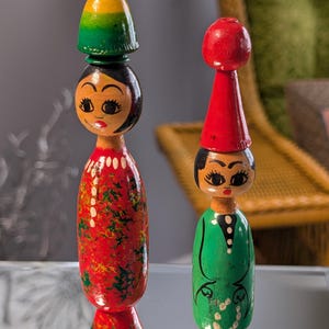 May include: Two hand-painted wooden figurines. One is red with green accents, a black head, and a yellow and green top. The other is green with a black head and a red top. Both have painted faces and are standing upright.