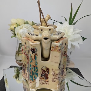 May include: A beige ceramic teapot with a square-shaped body and a lid featuring a central spout. The exterior is decorated with colorful illustrations of figures in traditional attire and landscape elements. A thin brown wire handle is attached.