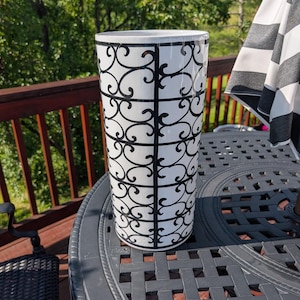 May include: A tall, cylindrical white vase with a black scrollwork design. The vase is made of ceramic and is approximately 12 inches tall. It is sitting on a dark gray outdoor table. The background includes a wooden deck, trees, and a black and white striped umbrella.