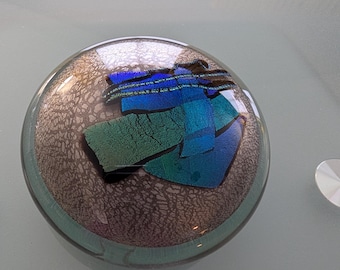 May include: A circular glass paperweight featuring a textured, metallic bronze-colored base layer with a web-like pattern. Embedded within the clear glass top are several overlapping, rectangular shards of iridescent blue and green dichroic glass.