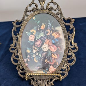 May include: An oval picture frame crafted from antique-toned brass with intricate scrollwork and filigree detailing. The frame holds a print depicting a vibrant floral arrangement featuring various colorful blossoms against a dark background.