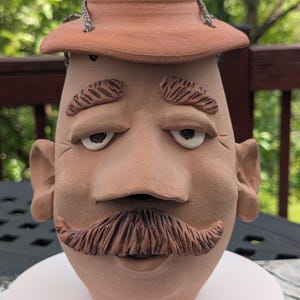 May include: A terracotta clay vessel shaped like a human head with a wide-brimmed hat, prominent ears, textured eyebrows, and a sculpted mustache. The vessel features a smooth, matte finish in a natural reddish-brown earth tone color.