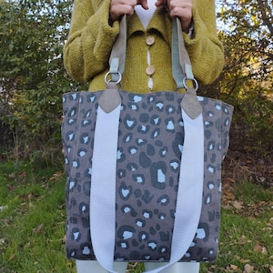 May include: A grey tote bag featuring a leopard print design in black and light blue. The bag has light blue straps and a brown handle. The bag is held by a person wearing a green cardigan.