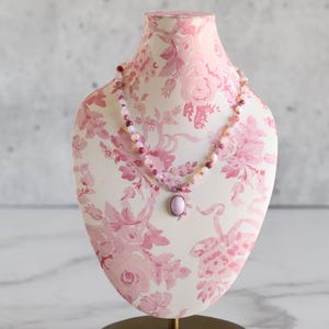 May include: A pink beaded necklace with a matching oval pendant is displayed on a floral print jewelry bust. The bust is white with a pink floral pattern and sits on a gold-colored base. The necklace features various shades of pink beads.