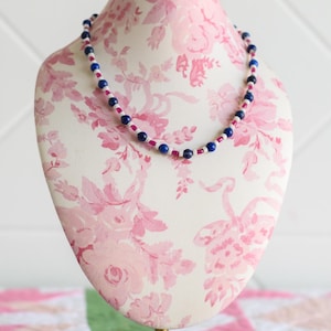 May include: A beaded necklace featuring a repeating pattern of round dark blue beads, small white cylindrical beads, and small translucent pink cylindrical beads. The necklace is displayed on a mannequin bust to show the circular shape of the strand.