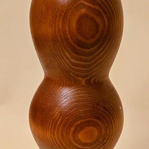 May include: A handcrafted wooden vase with a warm, reddish-brown hue. The vase has a unique hourglass shape, showcasing the natural wood grain patterns. The base is wider, narrowing in the middle, and widening again at the top.