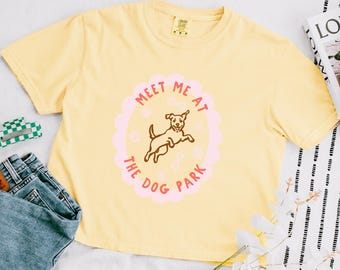 May include: Yellow t-shirt with a crew neck and short sleeves. The shirt features a pink oval design with the text "Meet Me At The Dog Park" and a brown illustration of a dog jumping. A pair of blue jeans is visible in the image.