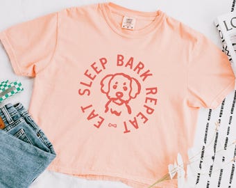 May include: Peach-colored t-shirt with a round graphic in coral featuring a dog illustration and the words "Eat, Sleep, Bark, Repeat." The shirt is short-sleeved and has a crew neck. The shirt is paired with blue jeans.