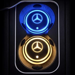 May include: A car interior cup holder with blue and yellow illuminated rings. Each ring features the Mercedes-Benz logo. The cup holder is set against a black background, with a sleek, modern design.