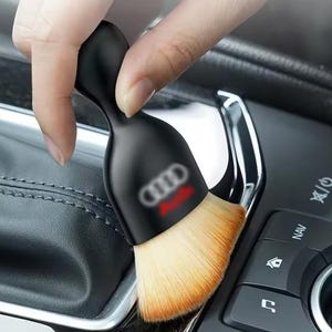 May include: A black car detailing brush featuring the Audi logo in red and white. The brush has a black handle and soft, light-coloured bristles. The brush is being used on a car's interior.