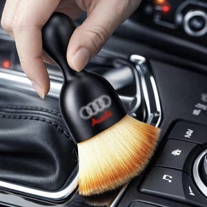 May include: A black car detailing brush with the Audi logo in white and red. The brush has a rounded handle and soft, light brown bristles. The brush is designed for cleaning car interiors.