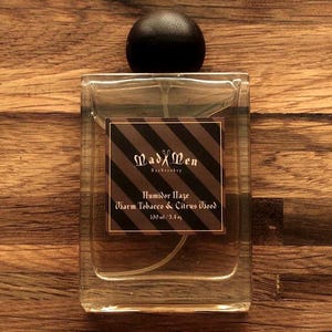 May include: A rectangular glass perfume bottle with a black wooden cap. The label features the text "MadXMen" and "Humidor Haze Warm Tobacco & Citrus Wood". The bottle contains 100 ml / 3.3 fl oz of fragrance.