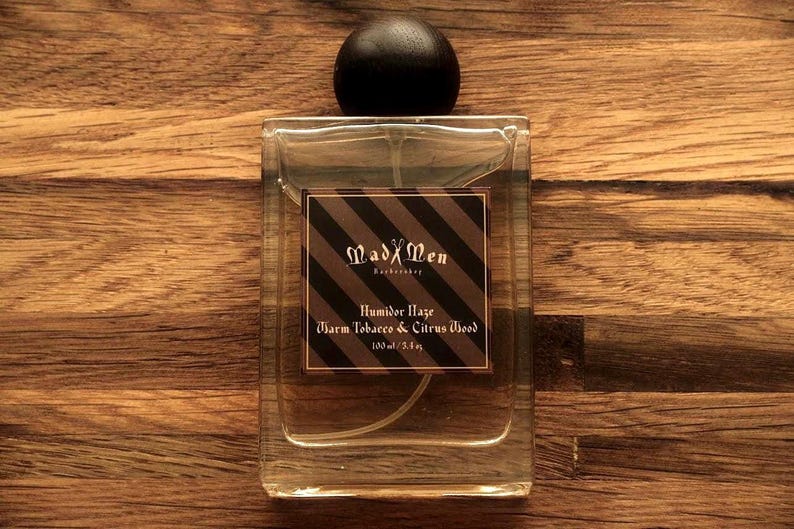 May include: A rectangular glass perfume bottle with a black wooden cap. The label features the text "MadXMen" and "Humidor Haze Warm Tobacco & Citrus Wood". The bottle contains 100 ml / 3.3 fl oz of fragrance.