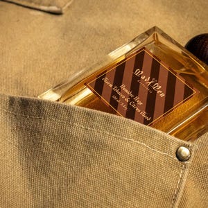 May include: A rectangular perfume bottle with a wooden cap is nestled in a tan pocket. The bottle has a brown and gold label that reads "Mad X Men Humidor Haze Warm Tobacco & Citrus Wood 3.4 fl oz". The pocket is part of a tan fabric.