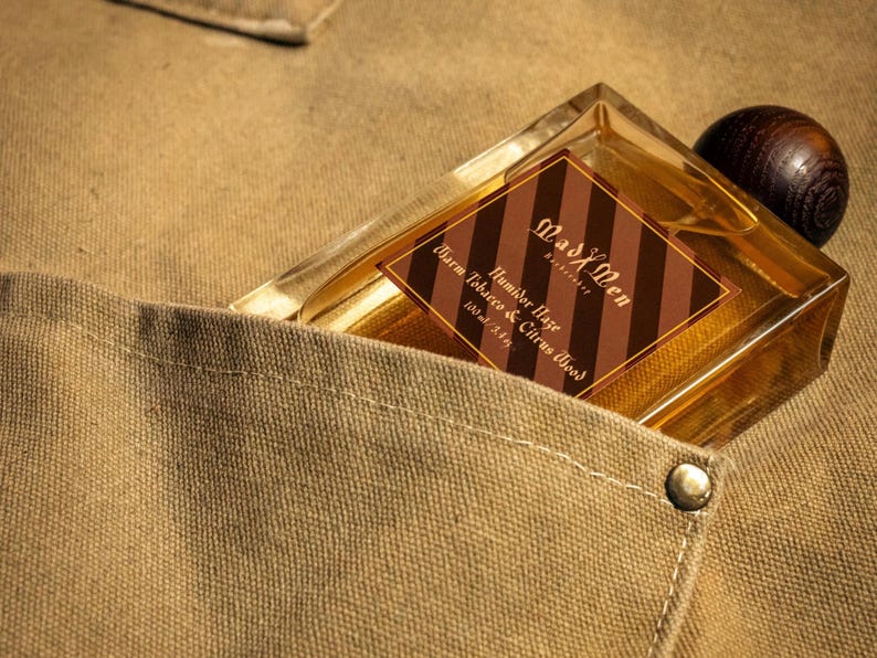 May include: A rectangular perfume bottle with a wooden cap is nestled in a tan pocket. The bottle has a brown and gold label that reads "Mad X Men Humidor Haze Warm Tobacco & Citrus Wood 3.4 fl oz". The pocket is part of a tan fabric.