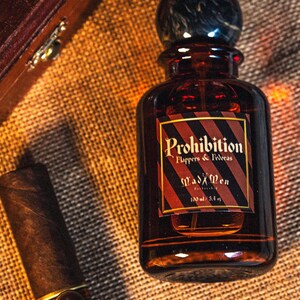 K&ouml;nnte beinhalten: Eine braune Glasflasche des Duftes "Prohibition" mit einem dunklen, marmor&auml;hnlichen Verschluss und einem Etikett mit der Aufschrift "Mad Men". Die Flasche ist 100 ml. Eine Zigarre liegt neben der Flasche auf einer strukturierten Oberfl&auml;che.